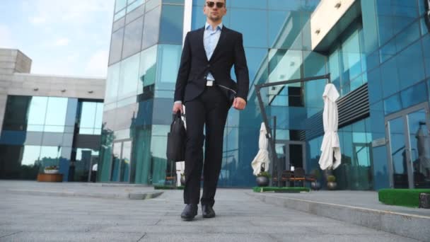 Close up de jovem empresário bonito em um terno preto com saco andando ao longo do edifício de escritórios moderno. Empreendedor do sexo masculino confiante com pasta vai trabalhar. Visão de ângulo baixo Movimento lento — Vídeo de Stock