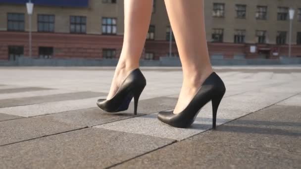 Slanke vrouwelijke benen in zwarte schoenen op hoge hakken lopen op het stadsplein op een zonnige dag. Voeten van jonge zakenvrouw in schoeisel op hoge hakken in stedelijke straat. Low angle view Slow motion Close up — Stockvideo