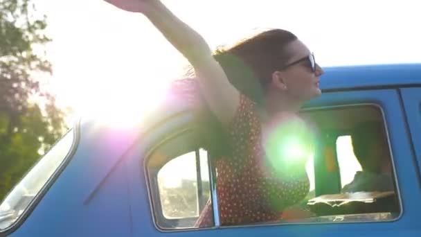 Młoda kobieta wyglądająca przez okno na poruszający się stary samochód. Szczęśliwa dziewczyna w okularach przeciwsłonecznych, wychylona przez okno samochodu i ciesząca się podróżą. Rozbłysk słońca w tle Koncepcja podróży i wolności. Powolny ruch Zamknij — Wideo stockowe