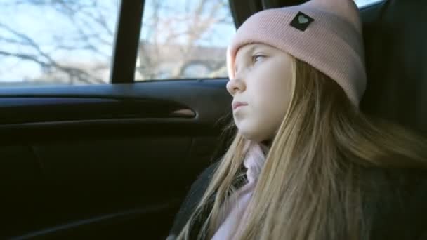 Hermosa niña con el pelo largo y rubio mira hacia fuera a través de la ventana en el asiento trasero de un coche en movimiento. Pequeña chica triste paseos en SUV moderno y relojes en la naturaleza a través del vaso de auto. Lento mo — Vídeos de Stock