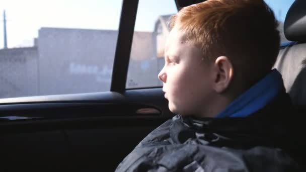 Close-up van de kleine mannelijke kind kijkt door het raam in de achterbank van een bewegende auto. Droevige roodharige jongen rijdt op het platteland op moderne SUV en kijkt naar de natuur door het glas van de auto — Stockvideo