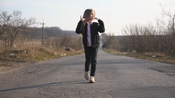 Blisko szczęśliwej uśmiechniętej dziewczyny w kurtce spaceruje po pustej drodze i zabawnie tańczy. Piękna kobieta z długimi blond włosami cieszy się i bawi na pustym podjeździe na wsi. Zwolniony ruch — Wideo stockowe