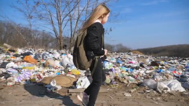 Egy kislány hátizsákkal megy az iskolába a szemétlerakó hátterében. Szomorú kislány szőke hajjal, sok szemét mellett sétál vidéken. Környezetszennyezés — Stock videók