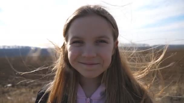 Primer plano de la niña pequeña y feliz mira a la cámara y endereza su cabello rubio que sopla sobre el fondo del campo arado. Retrato de niña alegre se para en el prado arado y ríe — Vídeos de Stock