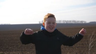 Küçük kızıl saçlı çilli çocuk kameraya bakar ve pullu çayırda ayakta alkışlayarak el kaldırır. Tarlada yüzü olumlu duygularla dolu mutlu bir erkek çocuğun portresi.