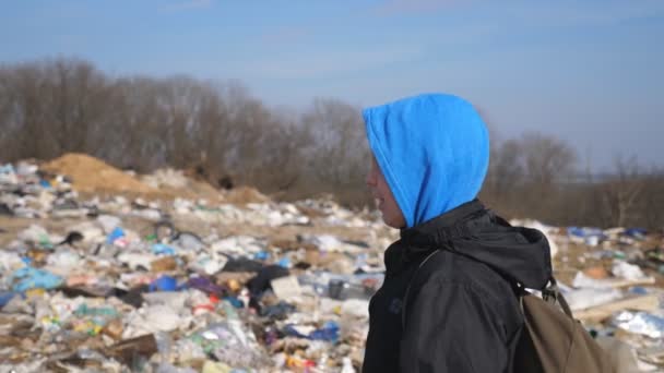 Close-up van kleine droevige jongen met rugzak loopt langs de weg naar school tegen de wazige achtergrond van vuilnisbelt. Klein mannelijk kind komt in de buurt van veel vuilnis op het platteland. Milieuverontreiniging — Stockvideo