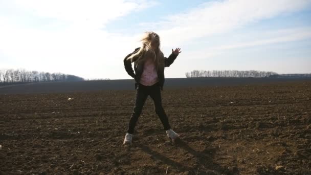 Schattig vrouwelijk kind met lang blond haar danst grappig op geploegd veld op zonnige dag. Close-up van gelukkig lachend meisje in jas geniet en heeft plezier op geploegde weide in het vroege voorjaar. Dolly schot — Stockvideo