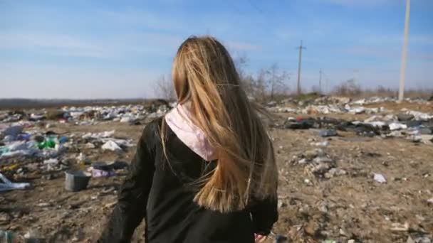 Close up de criança pequena fêmea com cabelo loiro longo caminha no depósito de lixo sujo. A menina vai ao lixo no ferro-velho. Conceito de problema de poluição ambiental. Visão traseira Movimento lento — Vídeo de Stock