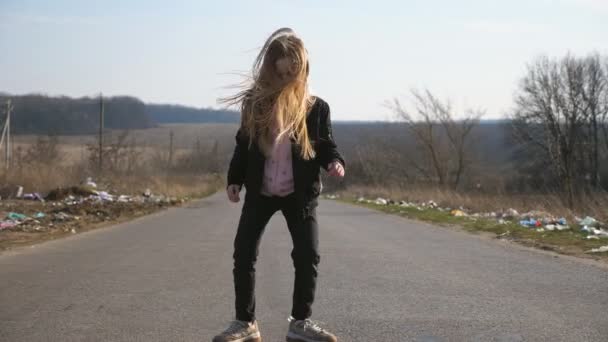 Bambino femminile allegro con lunghi capelli biondi balla divertente sulla strada in campagna. Primo piano di felice ragazza sorridente in giacca gode e si diverte sul vialetto vuoto nella giornata di sole. Rallentatore — Video Stock