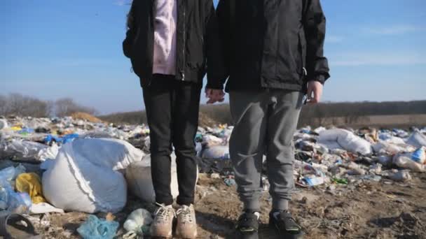 Retrato de una niña pequeña y un niño mira a la cámara contra el fondo del basurero. Los niños pequeños y serios se paran en el vertedero y se toman de la mano. Contaminación ambiental. Grúa tiro — Vídeos de Stock