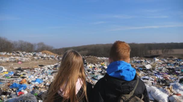 Die Rückansicht junger Mädchen und Jungen steht vor dem verschwommenen Hintergrund einer schmutzigen Müllhalde. Kleine Kinder betrachten Müll auf dem Schrottplatz. Konzept des Problems der Umweltverschmutzung. Nahaufnahme — Stockvideo