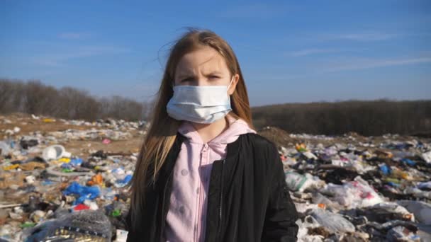 O retrato da pequena criança feminina na máscara protetora médica olha para a câmera contra o fundo do depósito de lixo sujo. A menina triste está no ferro-velho no campo. Poluição ambiental — Vídeo de Stock