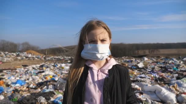 O retrato de uma menina séria na máscara protetora médica está no fundo da lixeira suja e olha para a câmera. Conceito de problema de poluição ambiental. Dolly atirou — Vídeo de Stock