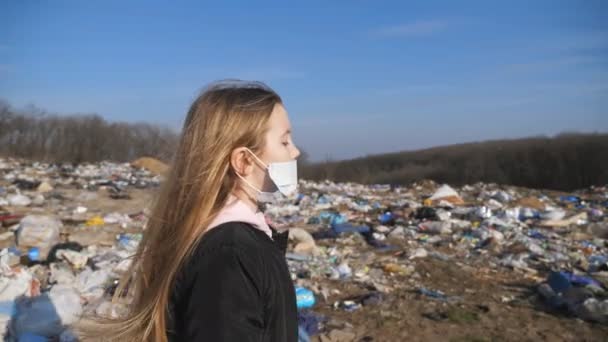 Pequena criança feminina em máscara protetora médica vai ao longo da estrada contra o fundo do depósito de lixo. Menina com cabelo loiro anda perto de um monte de lixo no campo. Poluição ambiental — Vídeo de Stock