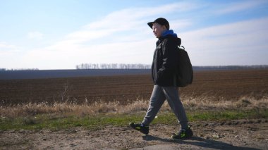 Ceketli mutlu erkek çocuğa yakın çekim, sürülen tarlanın yakınındaki bir garaj yolunda yapılır. Şapkalı küçük bir çocuk otlağın yakınındaki yolda yürüyor. Sırt çantalı küçük çocuk okula gidiyor..