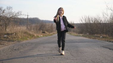 Ceketli mutlu gülümseyen kızın boş yolda yürümesi ve komik dans etmesi. Uzun sarı saçlı güzel bir bayan çocuk kırsal alanda boş bir garaj yolunda eğleniyor. Yavaş çekim.