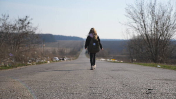 Small girl with backpack walks among road to training. Female child in jacket goes along a driveway to school. Unrecognizable little kid moves on way at sunny day. Blurred motion. Crane shot Slow mo.