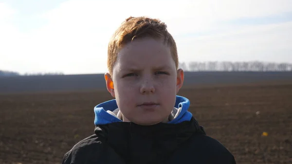 深刻な赤髪の男の子のカメラに耕起フィールドのぼやけた背景に見えます そばかすを持つ小さな子供は ぬかるんだ牧草地に立っています 顔に悲しい表情の男性の子供の感情を閉じます — ストック写真