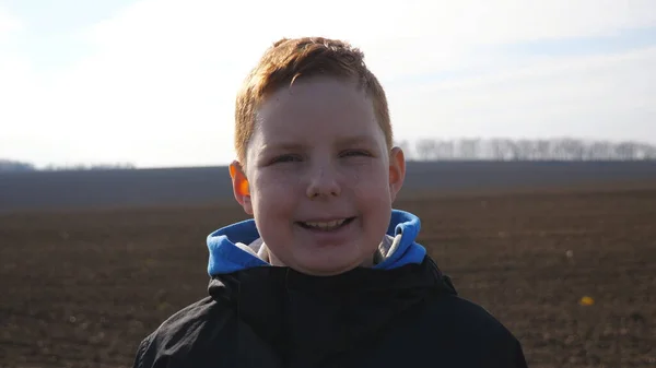 一个长着雀斑的有趣的生姜男孩的画像 在被犁地模糊的背景下看着摄像机 快乐的红头发小孩儿笑着站在犁地草地上 靠近点 — 图库照片