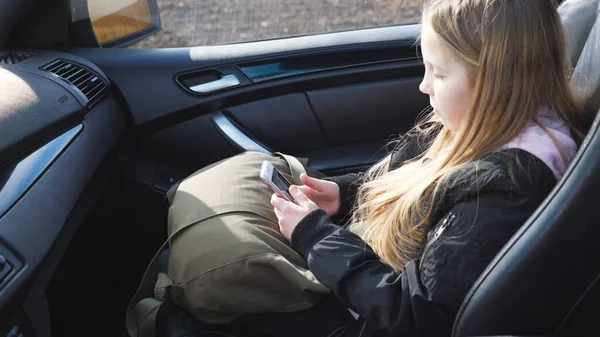 女の子は車の助手席に座って携帯電話でソーシャルメディアを閲覧します 長いブロンドの髪を持つ美しい女性の子供は 現代のSuvにあり 彼女のスマートフォンを使用しています スローモーション — ストック写真