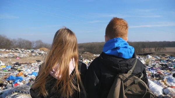 年轻的女孩和男孩站在肮脏的垃圾堆的模糊背景下 小孩子们手牵着手 看着垃圾场上的垃圾 环境污染的概念 — 图库照片