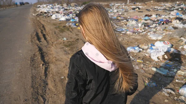 小女孩在垃圾堆的背景下上路了 一个金发的小女孩在乡间的车道上走着 旁边有很多垃圾 环境污染问题 — 图库照片