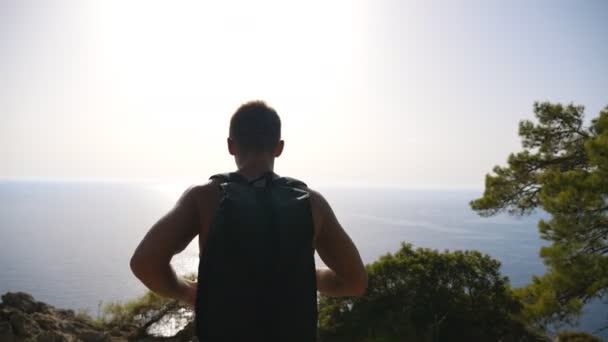 Caminante con mochila que llega hasta el borde de la montaña y levanta las manos admirando el paisaje marino. Hombre descansando activamente durante el viaje disfrutando de la libertad en la cima de la colina. Hermosa vista de fondo. Lento. — Vídeos de Stock