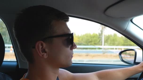 Bonito tipo de óculos de sol a conduzir a sua auto através da estrada rural no dia de verão. Perfil do homem cuidadosamente olhando para o tráfego e controlar seu veículo. Conceito de viagem de carro. Vista de dentro do carro — Vídeo de Stock