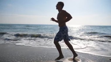 Güzel deniz manzarası arka planında koşan atletik yakışıklı adam. Okyanus sahilinde koşan genç sporcu. Erkek sporcu deniz kıyısında spor yapıyor. Sağlıklı yaşam tarzı kavramı. Yavaş çekim kenar görünümü