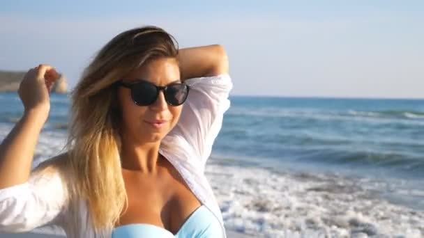 Vacker dam i solglasögon leende och leker med håret promenader längs kusten. Porträtt av kvinna njuter av frihet och ha kul på stranden under sommarlovet. Söt flicka återskapa på resort — Stockvideo