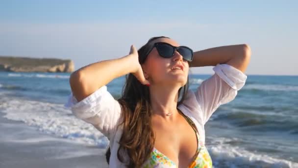 Gyönyörű hölgy napszemüvegben mosolyog és játszik a hajával sétált a parton. Portré nő élvezi a szabadságot és szórakozás a parton a nyári vakáció alatt. Aranyos lány újrateremtés üdülőhelyen — Stock videók