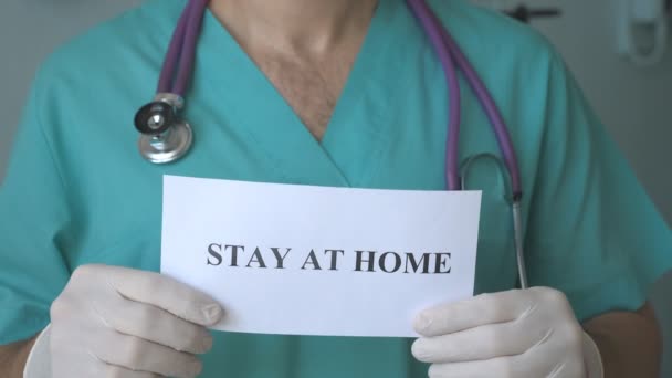 Médico masculino está segurando uma foto com a inscrição ficar em casa durante a epidemia de coronavírus. Médico jovem mostra cartaz com apelo ao auto-isolamento. Conceito de vida de segurança da pandemia de COVID-19 — Vídeo de Stock