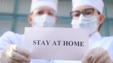Erkek ve kadın doktorlar, Coronavirus salgını sırasında evde kalan yazıtlı bir resim tutuyorlar. Sıhhiyeciler, COVID-19 salgınından kendini izole etme ve güvenli yaşam çağrısında bulunan bir poster gösteriyor.