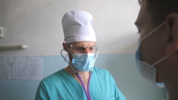 Doktor steteskoplu hastayı COVID-19 belirtileri için muayene ediyor. Sıhhiyeci insanın ciğerlerini dinler. Doktor, hastanedeki adamın göğsünü kontrol etti. Coronavirüs salgınından sağIık ve güvenlik kavramı — Stok video