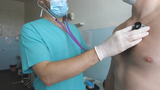 Médico con estetoscopio escucha los pulmones del paciente busca síntomas COVID-19. Doctor examinando al hombre con estetoscopio a los signos de neumonía por coronavirus. Médico revisando el pecho del tipo en el hospital — Vídeo de stock