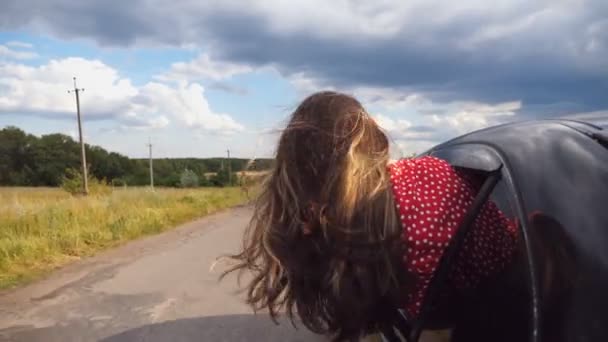 Brunette girl leaning out of car window and enjoying trip while riding through country road. Young woman sticking her head out of moving auto and her long brown hair blowing in wind. Close up Slow mo — Stock Video