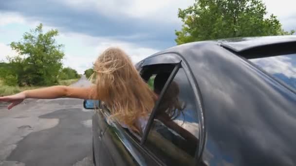 Klein meisje dat uit het autoraam leunt en haar hand uitsteekt terwijl ze door de landweg rijdt. Klein kind dat haar arm uit het open raam steekt en auto beweegt om de wind te voelen. Achteraanzicht Langzame beweging — Stockvideo