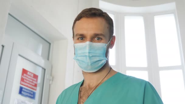 Portret van een jonge blanke arts met een medisch gezichtsmasker kijkt naar de camera. Medische werker draagt een beschermend masker tegen het virus. Concept van gezondheid en veiligheid van coronavirus pandemie. COVID-19 — Stockvideo