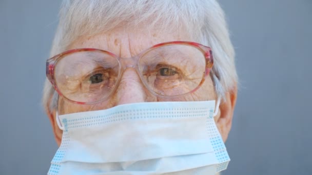 Portret van oma in bril draagt beschermende masker van het virus. Oma kijkt met scherp zicht in de camera. Concept van gezondheid en veiligheid als gevolg van een pandemie. Quarantaine van het coronavirus voor ouderen — Stockvideo