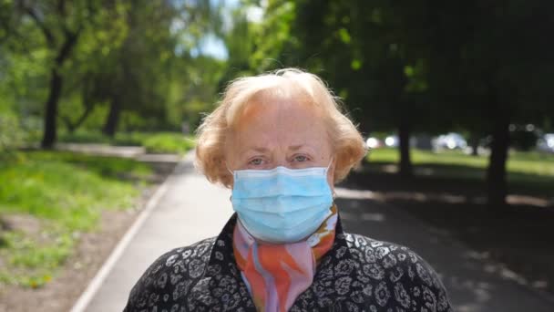 Na ulicy stoi portret staruszki w masce medycznej. Babcia zdejmuje maskę ochronną przed wirusem COVID-19 i poważnie patrzy w kamerę. Koncepcja bezpieczeństwa i higieny pracy koronawirusa — Wideo stockowe