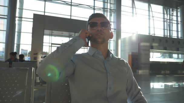 Jeune homme d'affaires souriant parlant au téléphone à l'aéroport avec éruption de soleil en arrière-plan. Gros plan portrait d'un homme d'affaires heureux et beau portant des lunettes de soleil et parlant sur son téléphone portable au bureau. Mouvement lent — Video