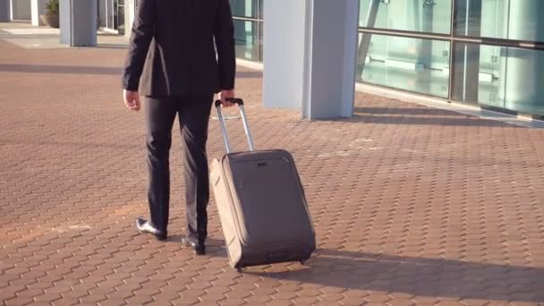 Vista posteriore di giovane uomo d'affari irriconoscibile in una tuta che cammina vicino al terminal e tira la valigia su ruote. Uomo d'affari di successo che va con i bagagli all'aeroporto. Chiudere rallentatore — Video Stock