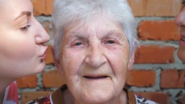 Genç kız ve yetişkin kadın sevgili büyükannelerini yanaklarından öpüyor. Gülümseyen ve kameraya bakan mutlu büyükannenin portresi. Sıcak aile ilişkileri. Sevgi ya da ilgi kavramı. Yavaş çekim