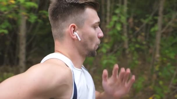 Atletico ragazzo sportivo con cuffie wireless che corre veloce lungo il sentiero vicino alla foresta. Giovane sportivo forte jogging all'aperto all'inizio dell'autunno. Un bell'uomo che corre verso la natura. Stile di vita sano e attivo — Video Stock