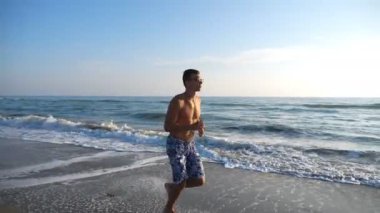 Atletik yakışıklı adam arka planda güzel bir deniz manzarasıyla sahilde koşuyor. Akşam vakti okyanus sahilinde koşan genç sporcu. Deniz kıyısında spor yapan erkek sporcu. Sağlıklı yaşam tarzı