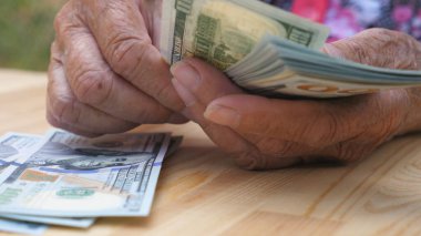 Buruşuk kadın ellerine düşük bir bakış açısı yabancı para birimini tutar ve masanın üzerinde nakit sayar. Yaşlı büyükannenin kolları masaya 100 dolarlık banknotlar koyar. Para konsepti. Yavaş çekim