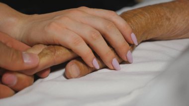 Torun ve torun, ninelerinin yatakta uzanan ellerini nazikçe okşuyor. Genç kadın ve erkek, klinikteki hasta büyükannenin kırışık kollarını rahatlatıyordu. İlgi ve sevgi kavramı. Kapat.