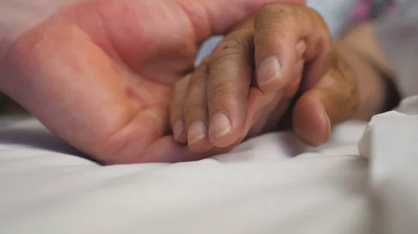 Young man holds and gently touches hand of sick mature woman lying in bed hospital. Grandson comforts wrinkled arm of his elderly grandmother in medical clinic. Son shows care and love to his parent.