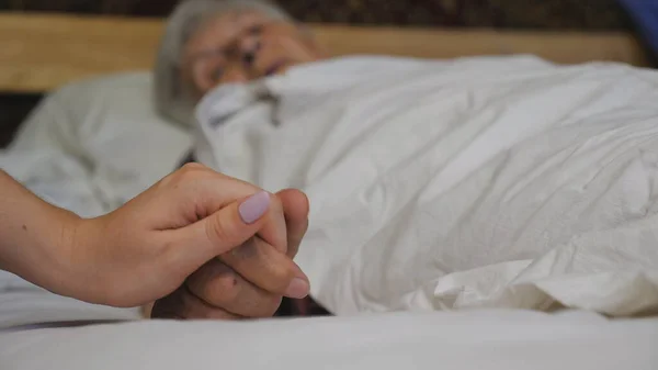 女孩抱着并轻柔地触摸躺在病床上的生病的成熟祖母的皱纹的胳膊 孙女在诊所里安慰她年老祖母的手 关心和爱的概念 靠近点 — 图库照片