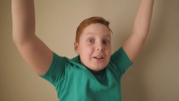 Ευτυχής αρσενικό παιδί τζίντζερ με φακίδες κοιτάζοντας στην κάμερα και σηκώνει τα χέρια χαρά επίτευγμα στο εσωτερικό. Πορτρέτο του μικρού κοκκινομάλλη αγόρι με θετικά συναισθήματα στο πρόσωπό του εσωτερικού χώρου. Αργή κίνηση — Αρχείο Βίντεο